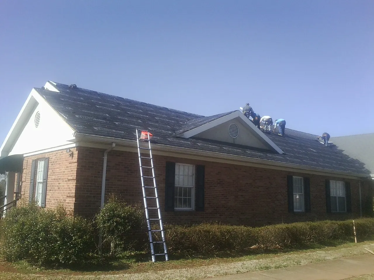 Crew tearing shingles off a brick ranch roof with a ladder set — Emergency Roof Repair work by our crew in Dryden