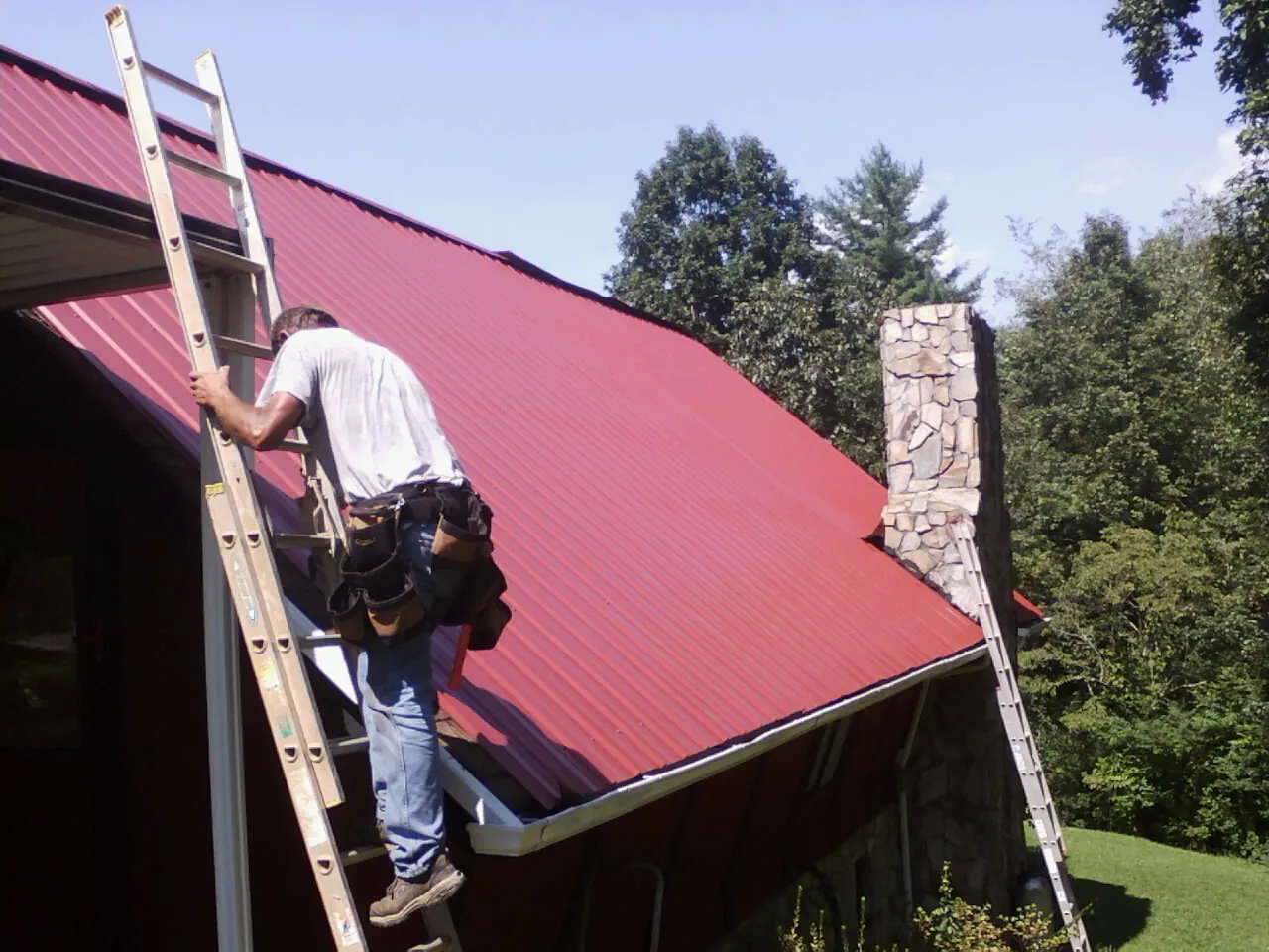 Roofer on a ladder beside a red metal roof and stone chimney — Soffit & Fascia Repair by Roofing Brothers in Dryden